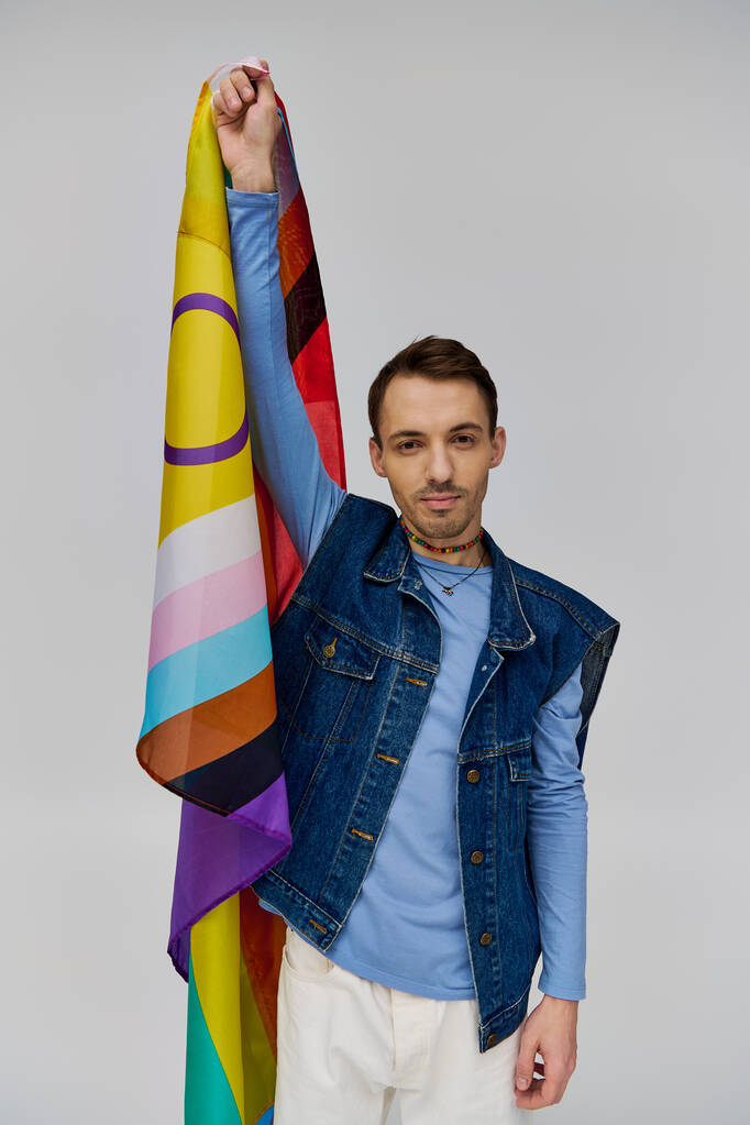 veselý pohledný gay muž v pulzující ležérní oblečení drží duhovou vlajku a usmívá se na kameru - Fotografie, Obrázek