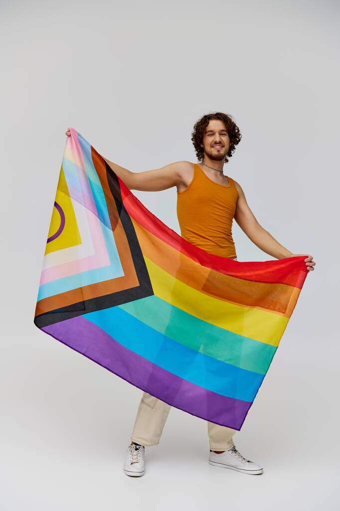 positivo seductor gay hombre con oscuro pelo sosteniendo arco iris bandera y sonriendo felizmente a cámara - Foto, imagen