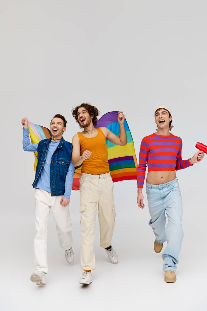 three trendy joyous gay men in cozy outfit holding rainbow flag and using megaphone on gray backdrop - Photo, Image