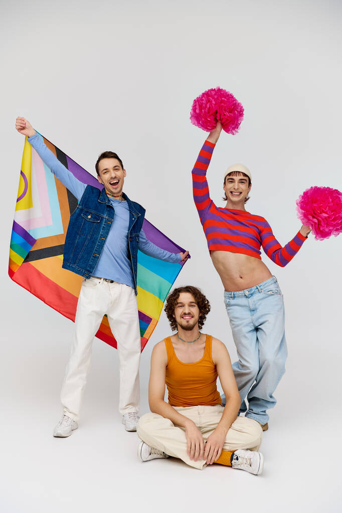alegre atractivo gay los hombres en vibrante ropa posando con arco iris bandera y pompones en gris telón de fondo - Foto, Imagen