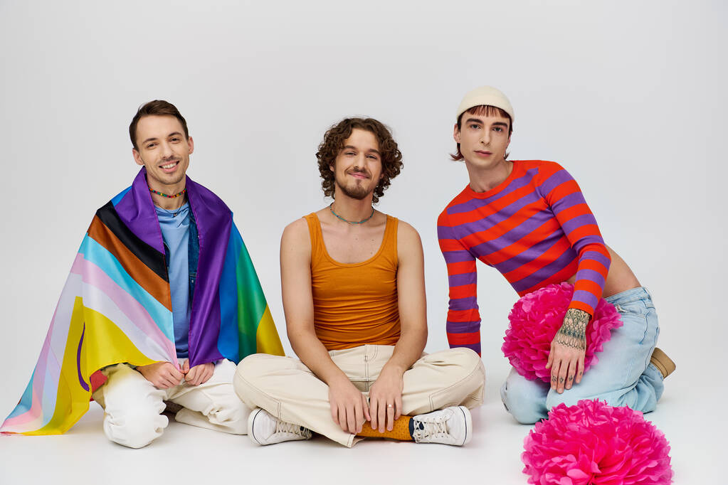 allegro attraente gay gli uomini in vibrante vestiti posa con arcobaleno bandiera e pon pon su grigio sfondo - Foto, immagini
