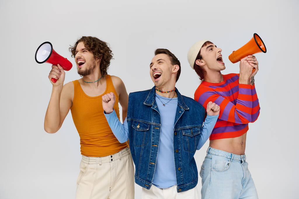 Три симпатичных весёлых гея в ярких одеждах, используя мегафоны, позируя на сером фоне - Фото, изображение