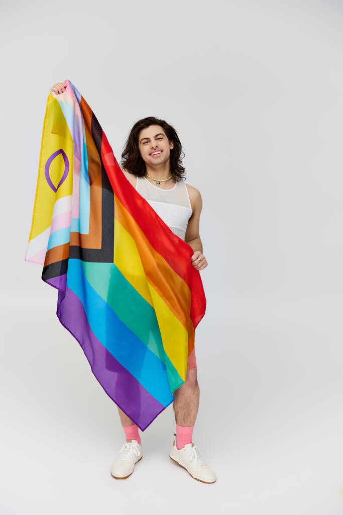 handsome joyous gay man with long dark hair posing with rainbow flag and looking at camera - Photo, Image