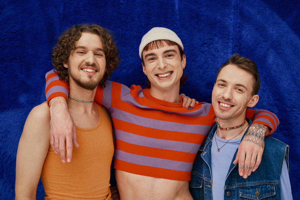 tres modish atractivo alegre gay amigos en acogedor atuendos posando en azul oscuro telón de fondo, orgullo mes - Foto, imagen