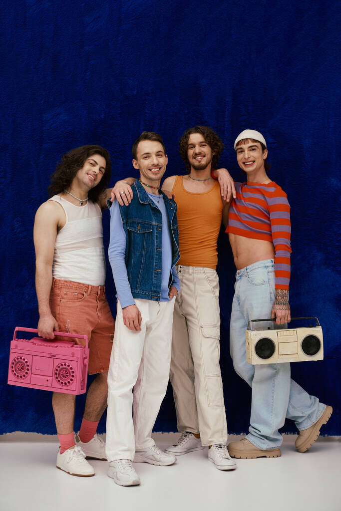 four joyful handsome stylish gay men in casual outfits posing with tape recorders, pride month - Photo, Image