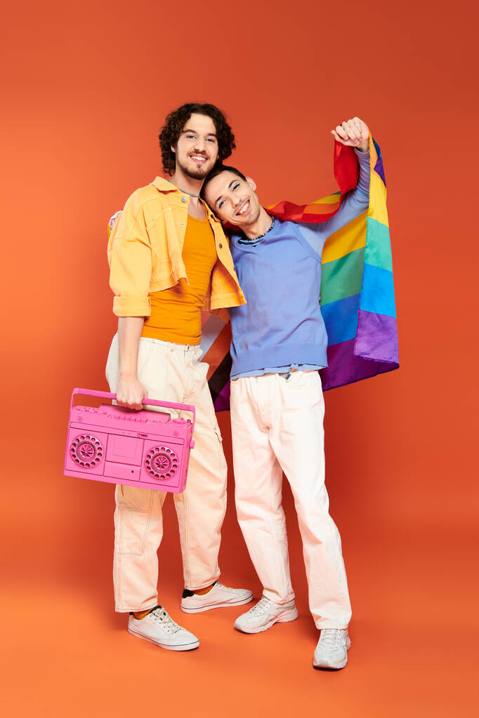 two positive handsome gay friends posing with tape recorder and rainbow flag on orange backdrop - Photo, Image