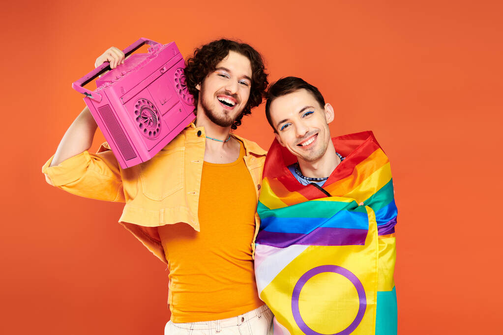 dos alegre guapo gay amigos posando con cinta grabadora y arco iris bandera en naranja telón de fondo - Foto, Imagen