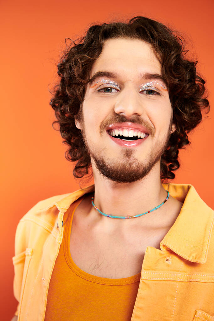 fröhlich ansprechender schwuler Mann mit dunklen Haaren und lebendigem Make-up posiert auf orangefarbenem Hintergrund, stolzer Monat - Foto, Bild
