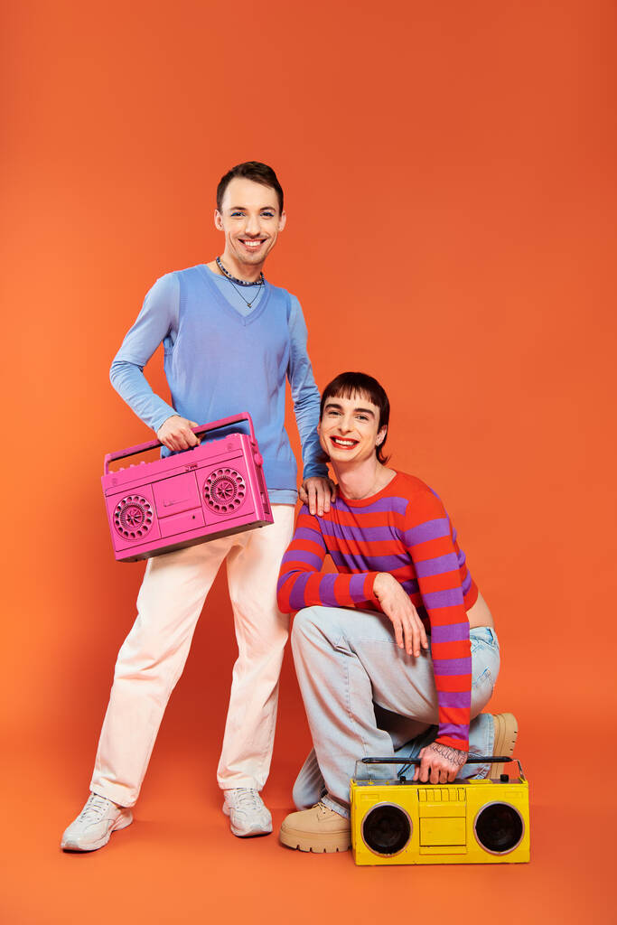 two joyful good looking gay men with vibrant makeup posing with tape recorders on orange backdrop - Photo, Image