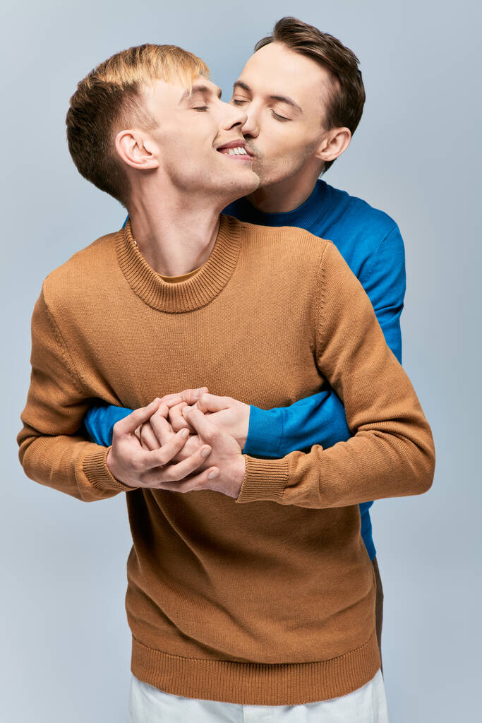 Двое мужчин в свитерах нежно целуются на сером фоне. - Фото, изображение