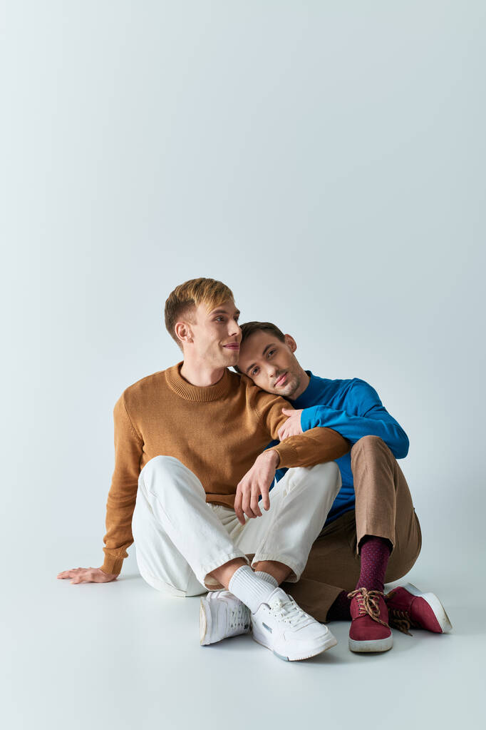 Dos hombres en atuendos casuales sentados en el suelo con los brazos alrededor el uno del otro en el fondo gris. - Foto, Imagen