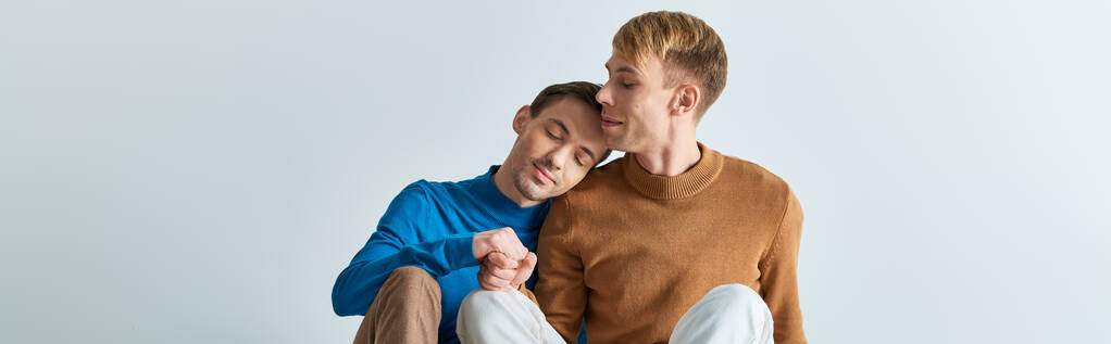 Zwei Männer in lässiger Kleidung, einer hält sein Bein hoch, teilen einen Moment der Intimität vor grauem Hintergrund.. - Foto, Bild