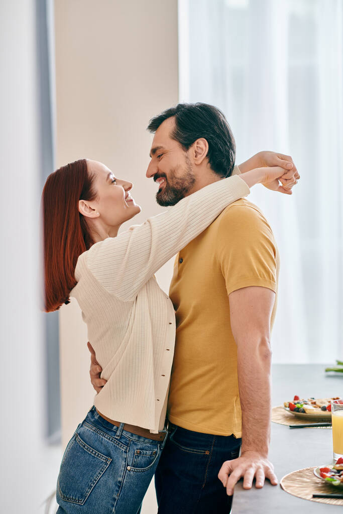 Dospělý pár, zrzka žena a vousatý muž, sdílejí něžné objetí ve své moderní kuchyni, vyjadřují lásku a blízkost. - Fotografie, Obrázek