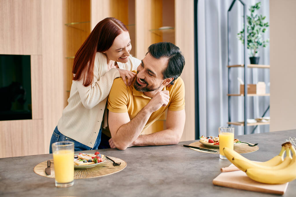 Egy gyönyörű vörös hajú nő és egy szakállas férfi együtt reggeliznek a modern konyhájukban.. - Fotó, kép