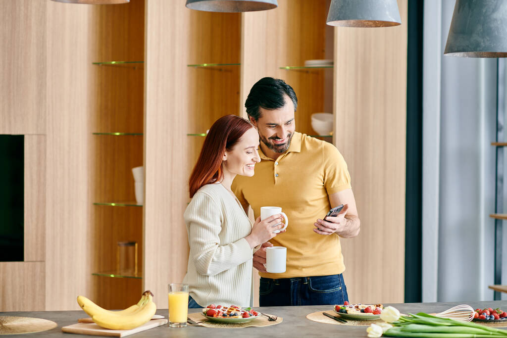 Een roodharige vrouw en een man met baard staren naar hun telefoon, staan in een moderne keuken, genieten van quality time thuis. - Foto, afbeelding