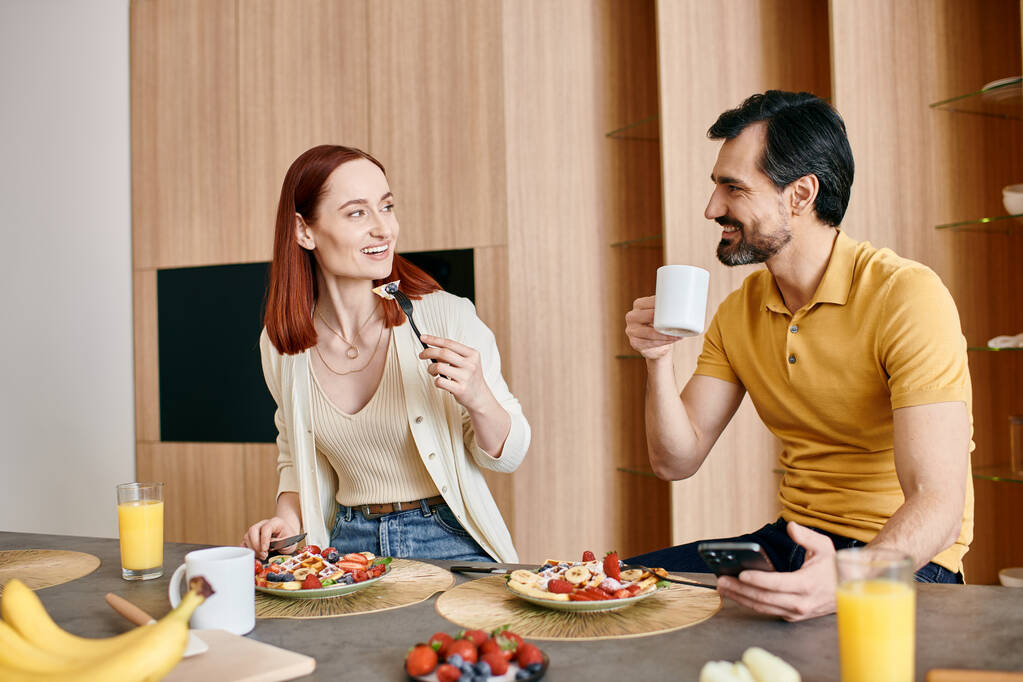 Uma mulher ruiva e um homem barbudo estão sentados em uma mesa de cozinha, comendo o café da manhã juntos em um apartamento moderno.. - Foto, Imagem