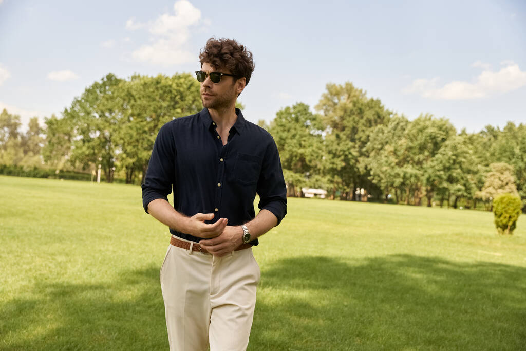 Un hombre con una camisa azul y pantalones bronceados disfruta de un paseo tranquilo en un exuberante campo verde bajo el vasto cielo abierto. - Foto, Imagen