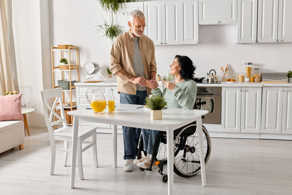 Niepełnosprawna kobieta na wózku inwalidzkim rozmawia z mężem w ciepłej atmosferze ich kuchni w domu. - Zdjęcie, obraz