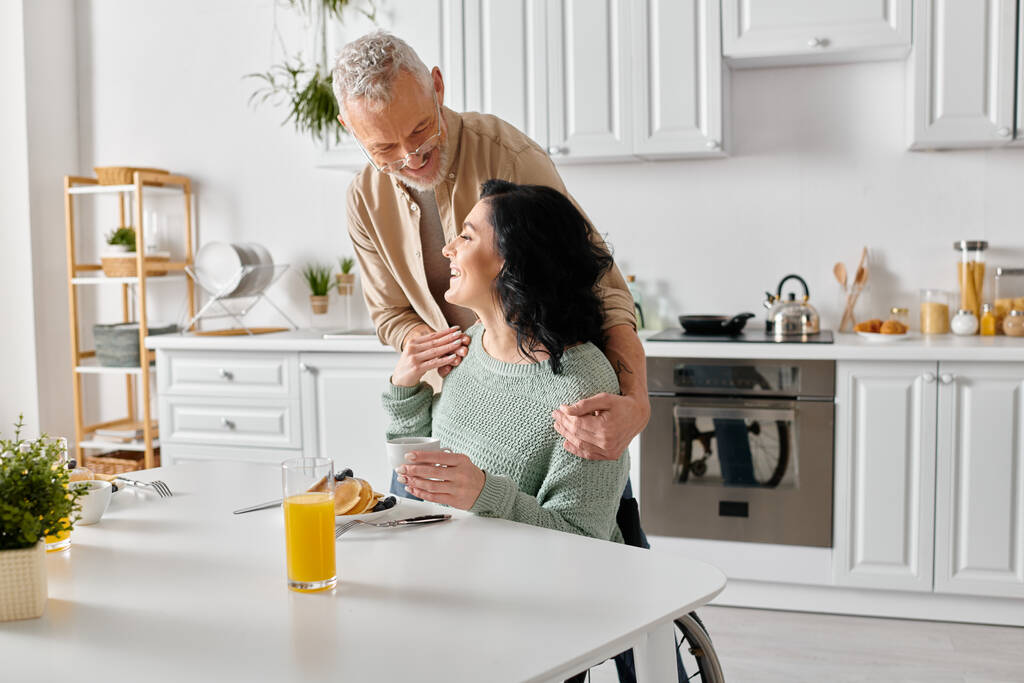 Manžel stojí vedle své postižené ženy na invalidním vozíku a tráví spolu chvilku v útulné kuchyni doma. - Fotografie, Obrázek
