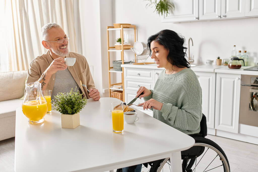 Una donna disabile in sedia a rotelle e suo marito condividono un momento caldo al tavolo della cucina nella loro casa. - Foto, immagini