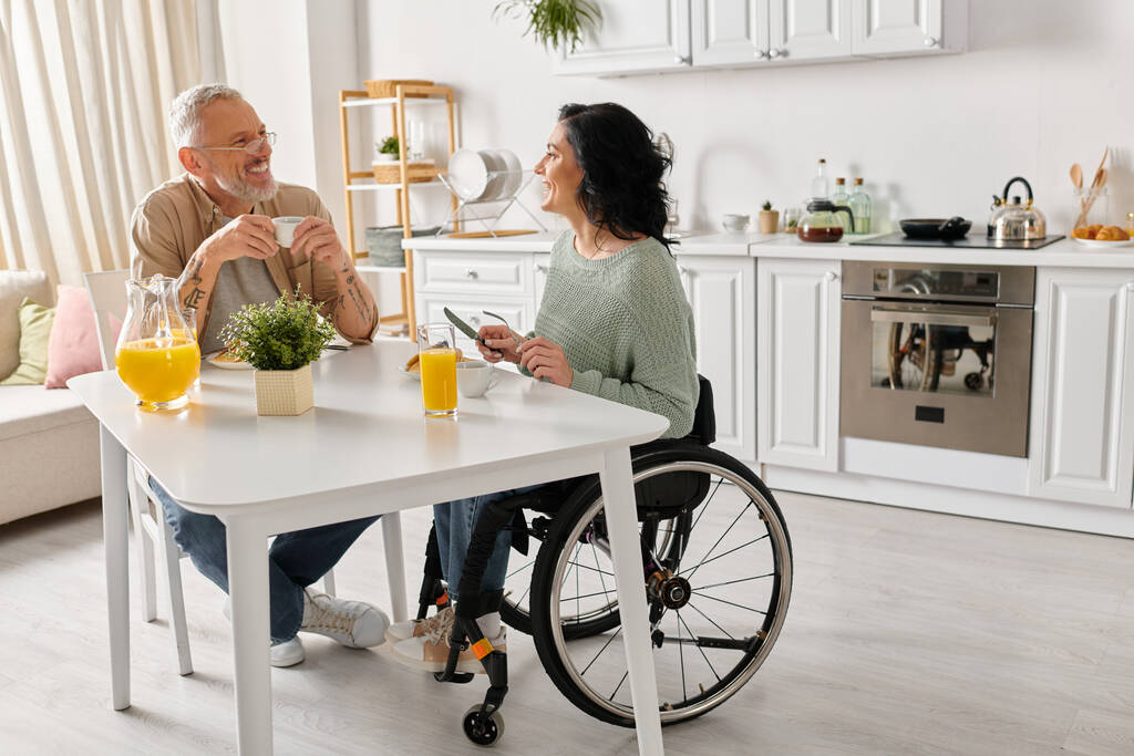 Un hombre en silla de ruedas conversando con una mujer en silla de ruedas en una acogedora cocina en casa. - Foto, imagen