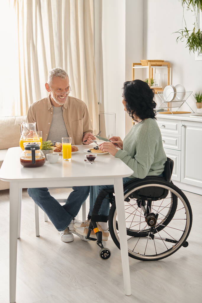 Un homme en fauteuil roulant et une femme à une table dans leur cuisine à la maison, partageant un moment de convivialité. - Photo, image