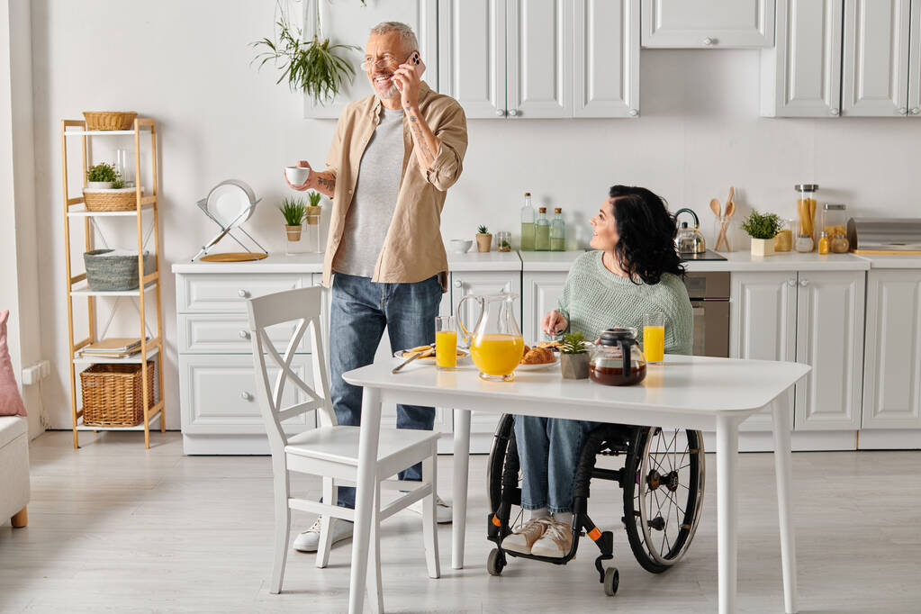 障害のある妻の横に立っている男性は,自宅のキッチンのテーブルに車椅子に座っています.. - 写真・画像