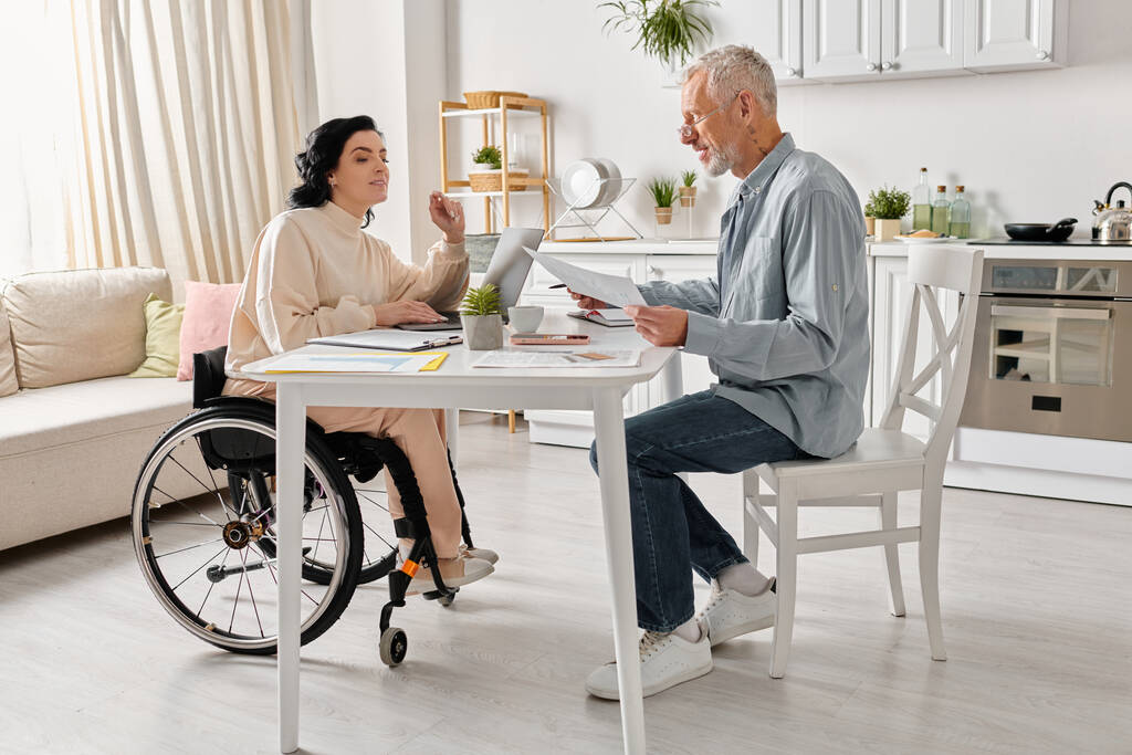 Ένας άντρας κάθεται σε ένα τραπέζι δίπλα σε μια γυναίκα σε αναπηρική καρέκλα στην κουζίνα τους στο σπίτι. - Φωτογραφία, εικόνα