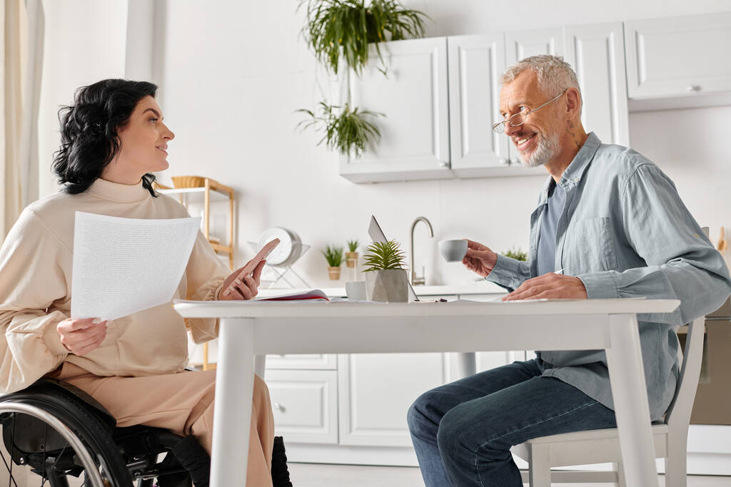 Un homme en fauteuil roulant engage une conversation avec une femme dans une cuisine à la maison. - Photo, image