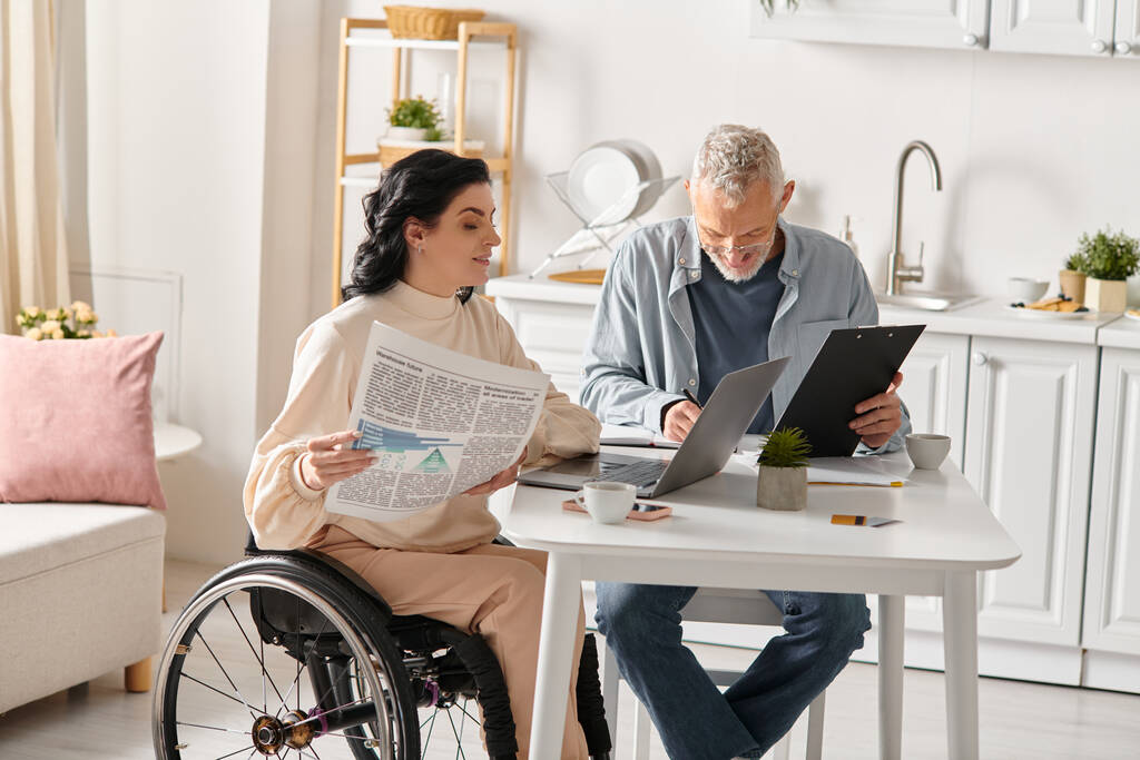 Un homme et une femme handicapée s'assoient à une table, se concentrant intensément sur un écran d'ordinateur portable dans leur cuisine confortable à la maison. - Photo, image