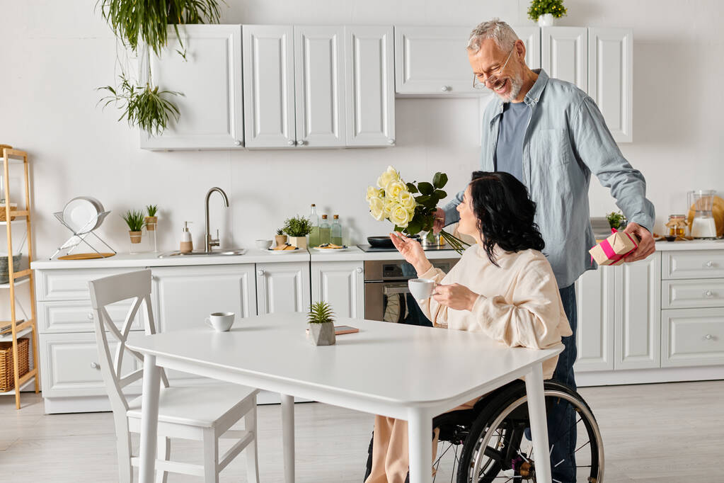 Мужчина ласково дарит букет цветов своей жене в инвалидном кресле на их кухне дома. - Фото, изображение