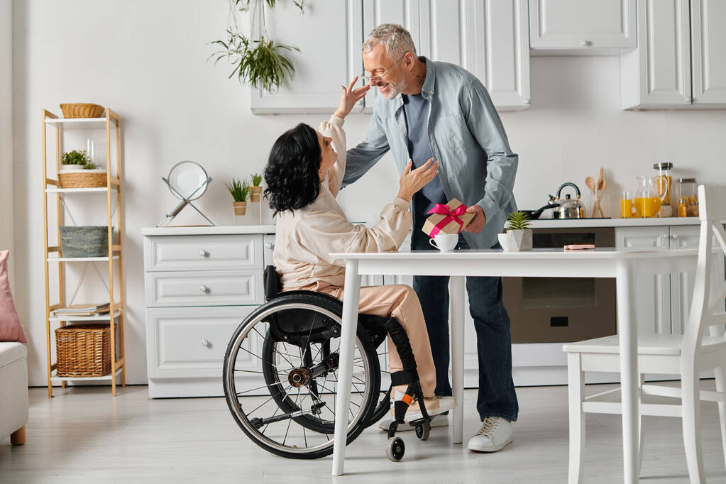 Un hombre cariñoso le da un regalo a su esposa feliz en una silla de ruedas, en la cocina de su casa. - Foto, imagen