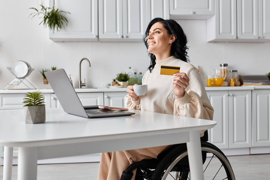 Uma mulher em uma cadeira de rodas confiantemente faz compras on-line com um cartão de crédito e laptop em sua cozinha. - Foto, Imagem