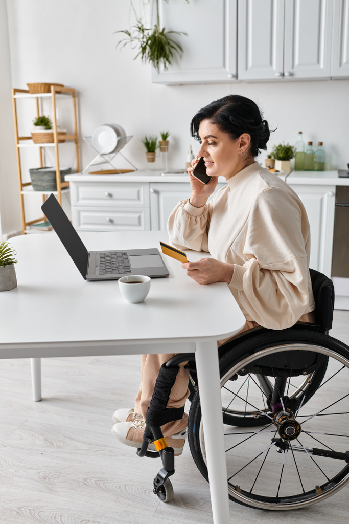 Uma mulher deficiente em uma cadeira de rodas trabalhando remotamente de sua cozinha, falando em um telefone celular. - Foto, Imagem