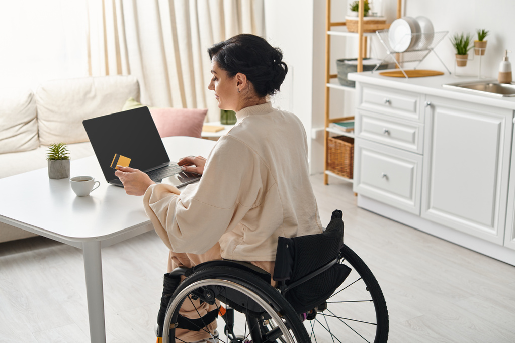 Una donna disabile su una sedia a rotelle sta lavorando a distanza sul suo portatile in cucina. - Foto, immagini