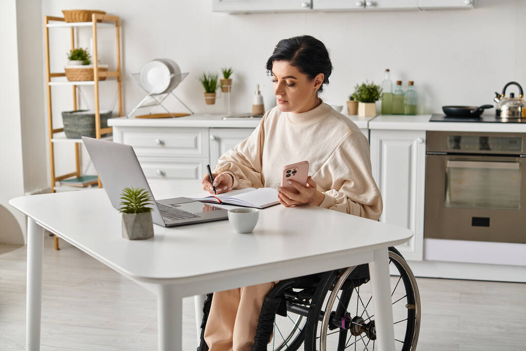 Een gehandicapte vrouw in een rolstoel met een laptop aan een keukentafel, die vastberaden en doelgericht op afstand werkt. - Foto, afbeelding