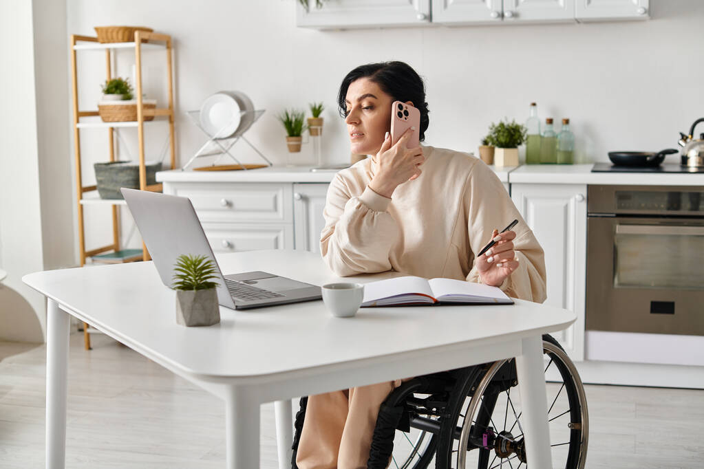 Mulher em cadeira de rodas trabalhando remotamente, conversando no celular na cozinha. - Foto, Imagem
