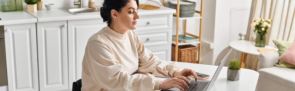 Uma mulher deficiente em uma cadeira de rodas, trabalhando remotamente, senta-se em uma mesa usando um computador portátil em sua cozinha. - Foto, Imagem