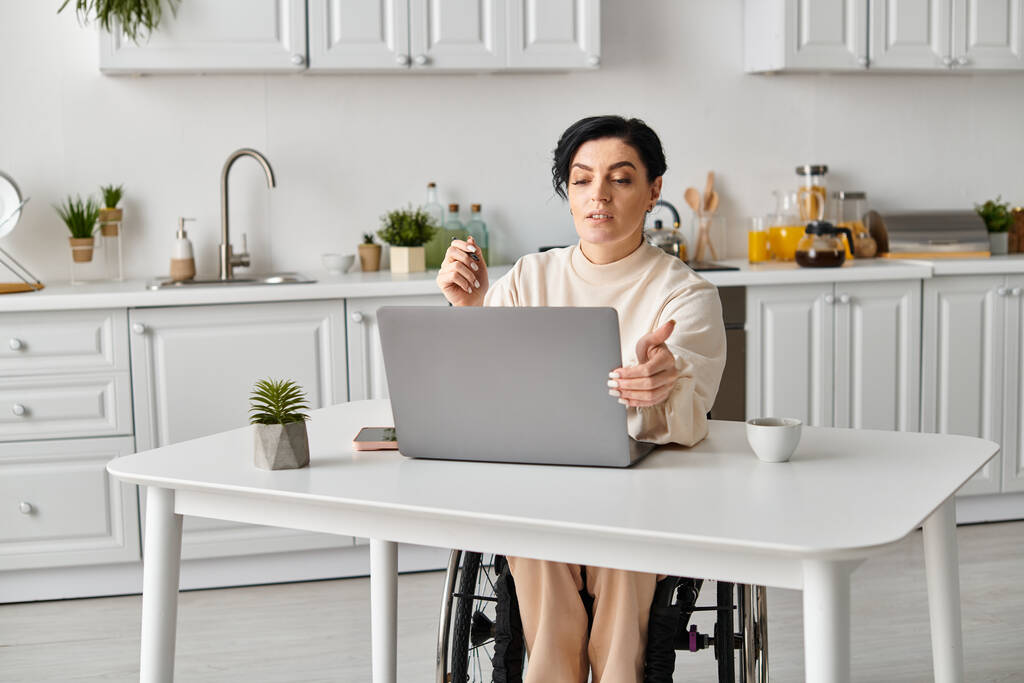 Uma mulher com deficiência em uma cadeira de rodas está focada em seu laptop em uma mesa de cozinha, participando de atividades remotas de trabalho ou lazer.. - Foto, Imagem