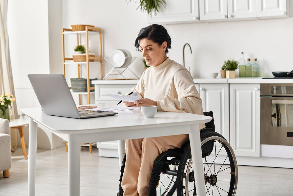 キッチンからラップトップで遠隔操作をしている車椅子の障害のある女性. - 写真・画像