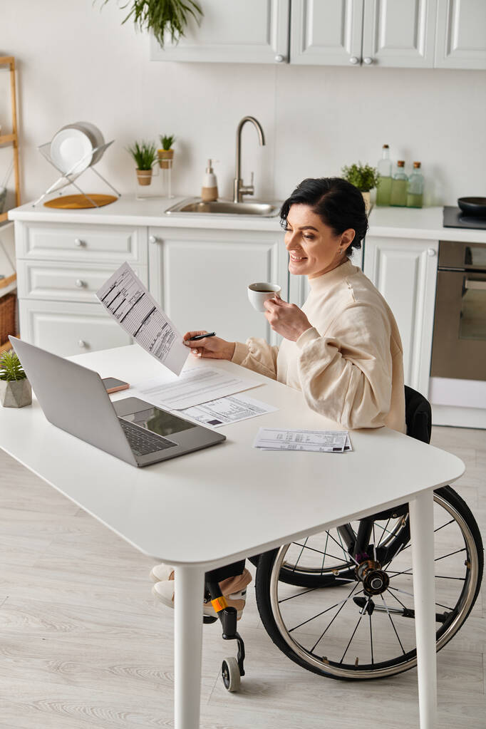 Kobieta na wózku inwalidzkim siedzi przy stole w kuchni, skupiona na zdalnej pracy na laptopie.. - Zdjęcie, obraz