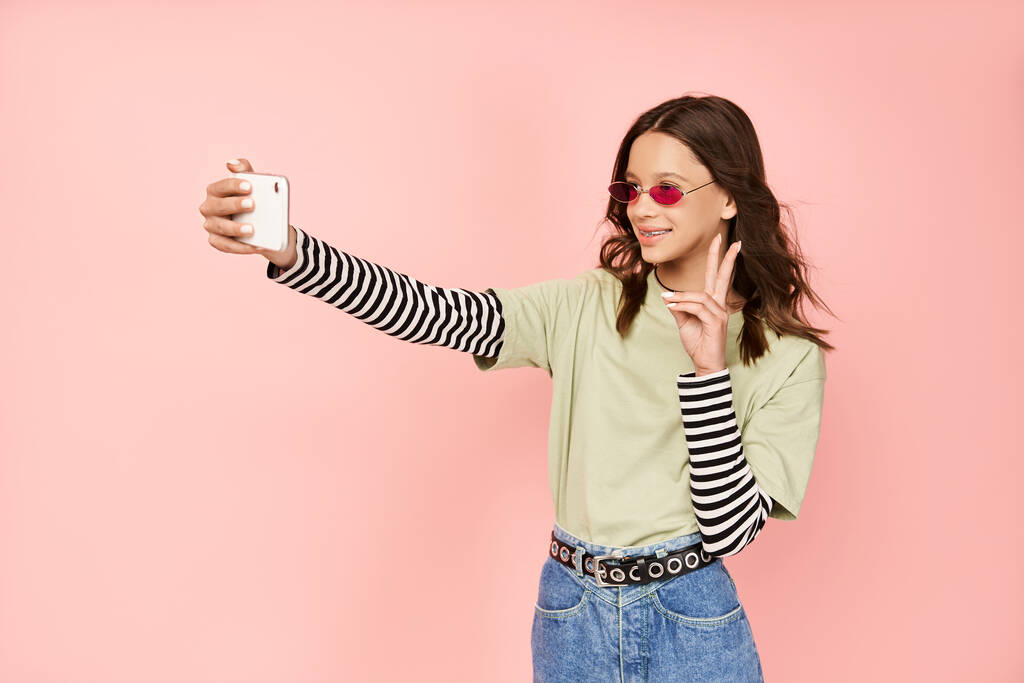 Een modieuze tiener meisje poseert zelfverzekerd, het vastleggen van een selfie met haar mobiele telefoon. - Foto, afbeelding