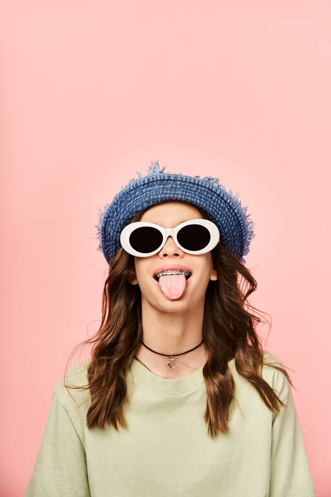 Egy elegáns tinédzser lány élénk ruhában vicces arcot vág napszemüvegben és kalapban.. - Fotó, kép