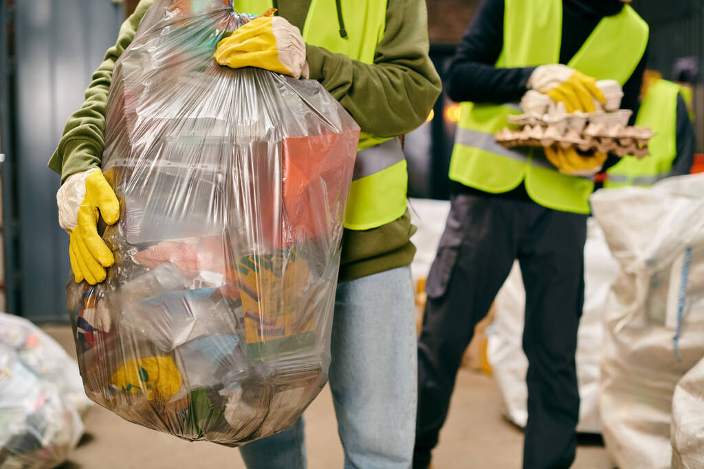 Two young volunteers in safety vests, gloves, and holding a huge bag of garbage while sorting trash. - Foto, imagen