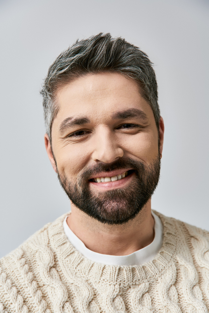 Een serene, grijsharige man met een baard, in een witte trui, glimlacht warm tegen een grijze studio achtergrond. - Foto, afbeelding