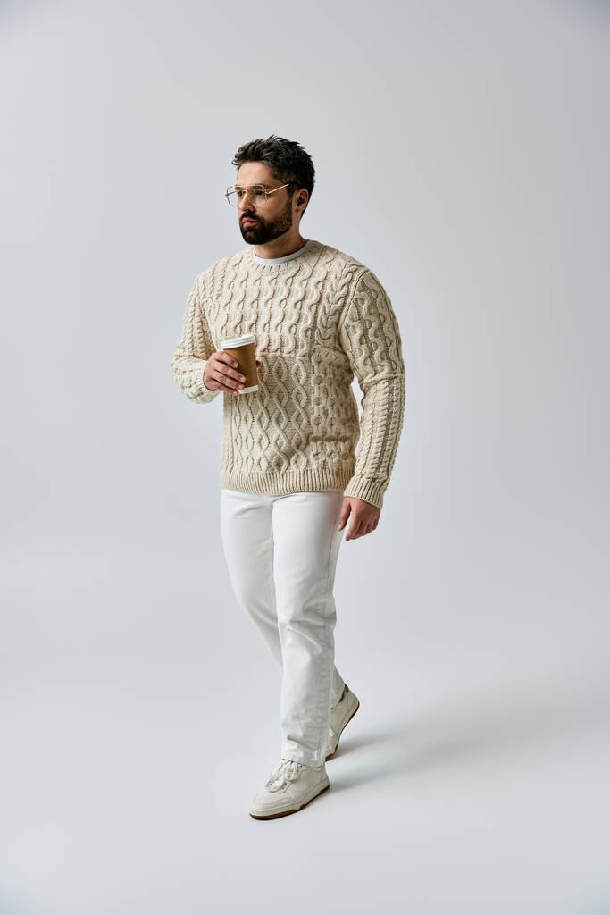 Ein bezaubernder Mann mit Bart posiert in weißem Pullover und passender Hose vor grauer Studiokulisse. - Foto, Bild