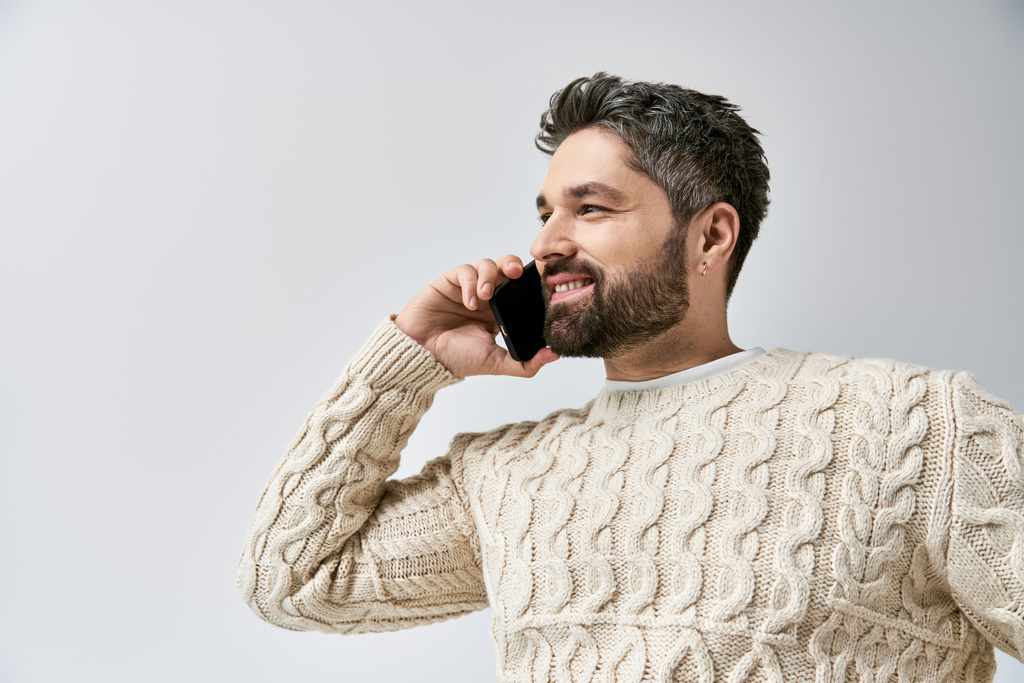 Egy divatos, szakállas férfi, aki mobilon beszél, miközben fehér pulóvert visel szürke háttérrel egy stúdióban.. - Fotó, kép