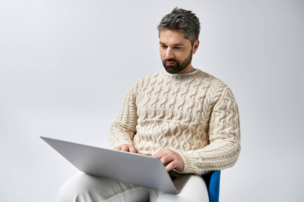 Un homme captivant avec une barbe absorbée par l'utilisation d'un ordinateur portable assis sur une chaise dans un contexte de studio gris. - Photo, image
