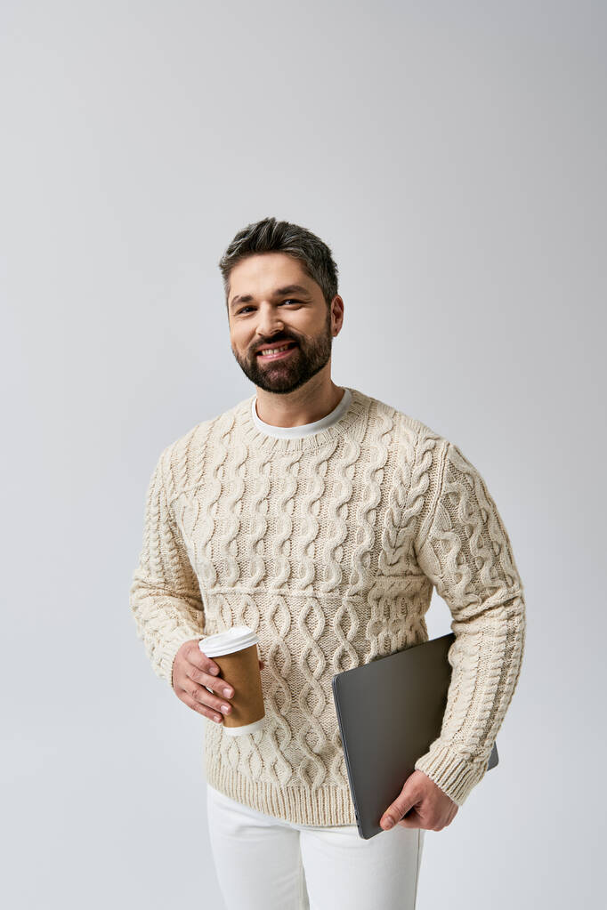 Een man met baard in een witte trui houdt een koffiebeker en een laptop tegen een grijze achtergrond. - Foto, afbeelding