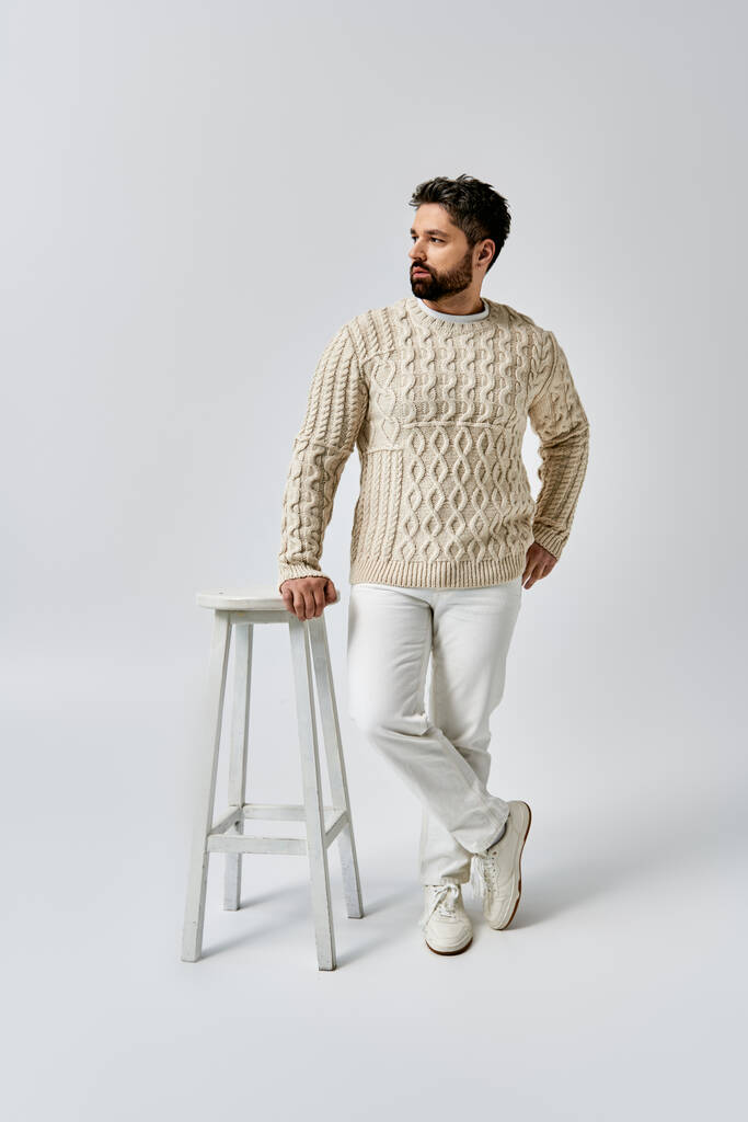 Egy szakállas, elegáns férfi magabiztosan áll egy szék mellett, divatos fehér pulóvert visel egy stúdióban.. - Fotó, kép